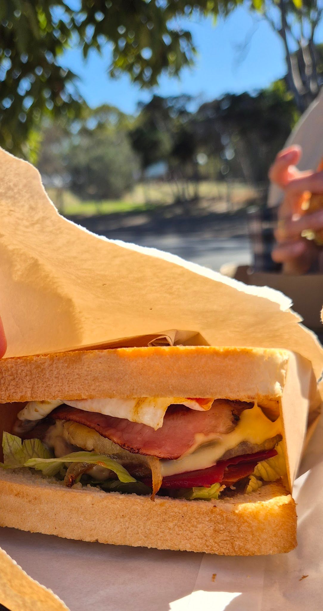 Overstuffed burger with egg, bacon, cheese, beetroot and lettuce held in brown paper outdoors
