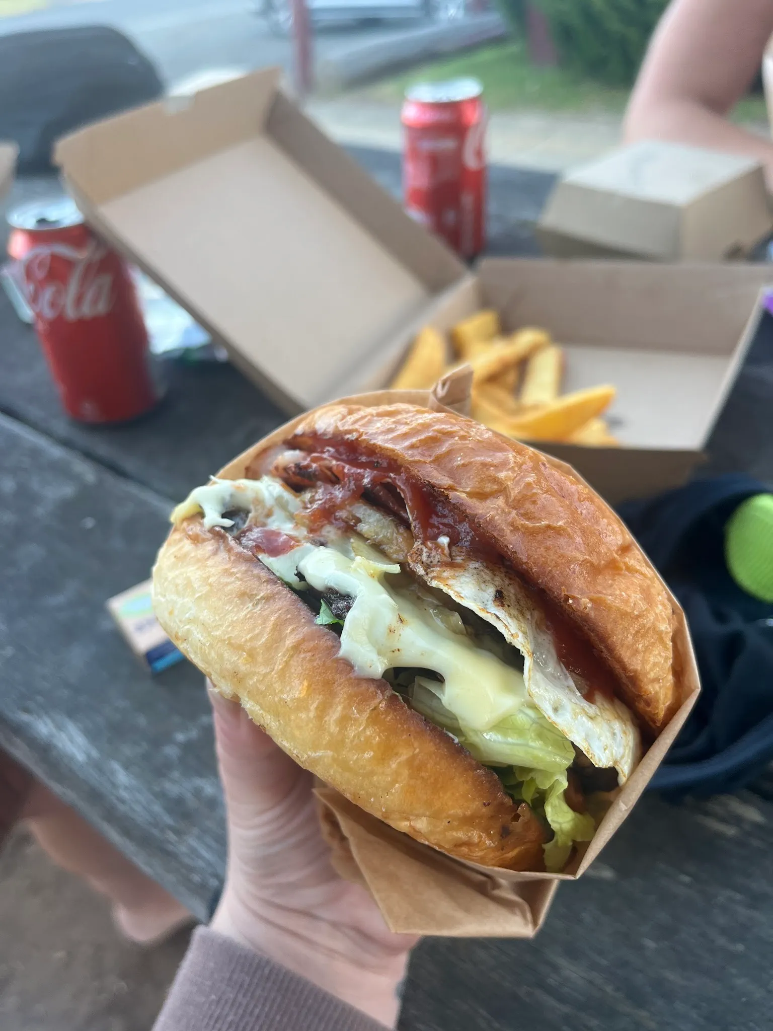 Large burger with the lot held in hand at a picnic table with chips and Coca-Cola in the background